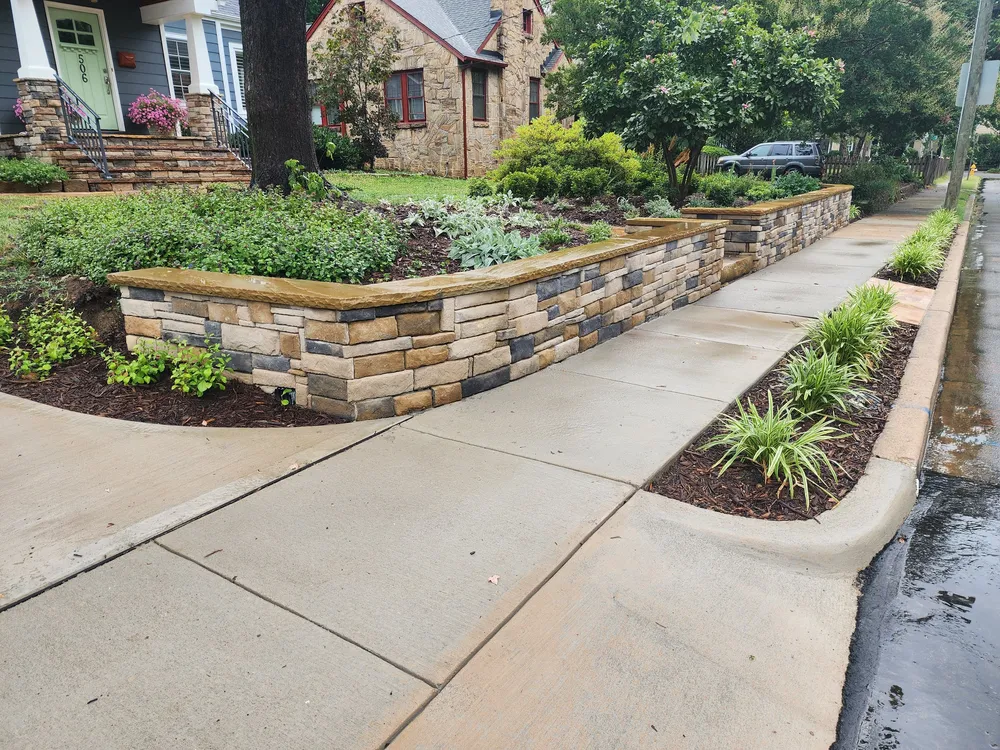 Stacked stone retaining wall with chiseled stone caps running along the sidewalk, with a planted bed behind it