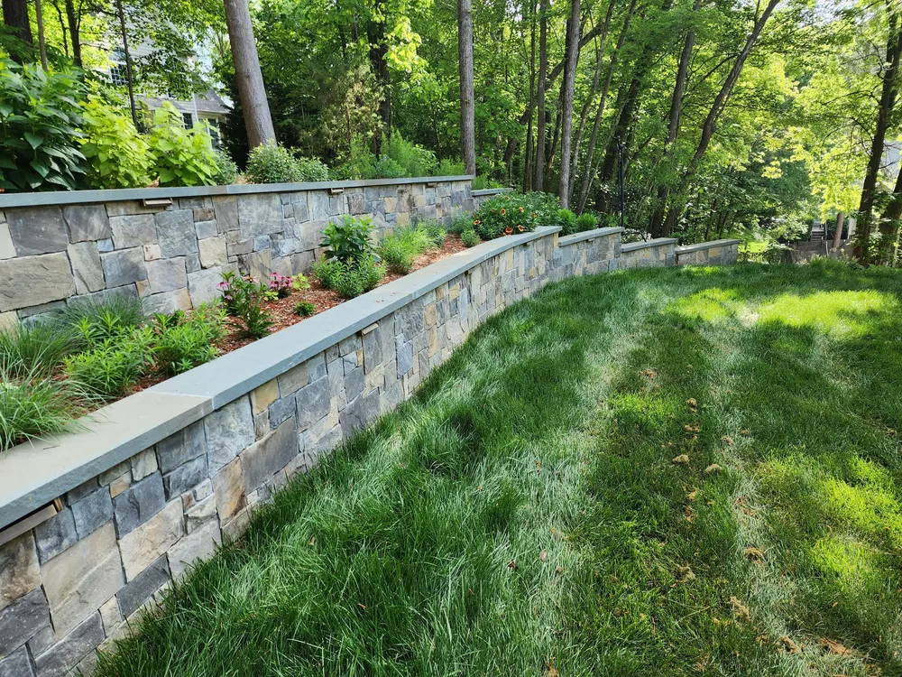 Two tiers of natural stone retaining walls with wall lights and perennial plantings above a curved lawn